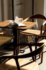 A cozy cafe setting with wooden furniture. An open book and a cold drink with a straw are placed on a sunlit table, surrounded by light-colored wooden chairs. The scene suggests a quiet and relaxing atmosphere.