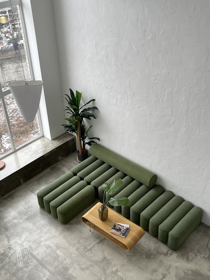 A modern living room space with a unique green modular sofa set against a textured white wall. The sofa consists of several cylindrical cushions arranged to form seating. In front of the sofa, there's a sleek, wooden coffee table with a vase holding green leaves and a couple of magazines on top. Next to the sofa is a tall potted plant with large leaves, adding a touch of greenery to the room. Light streams in through a large window, illuminating the concrete floor.