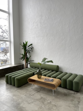 A modern living room with a green modular couch featuring cylindrical cushions and a sleek wooden coffee table with a vase containing leaves. The room has a tall potted plant in the corner and large windows allowing natural light to illuminate the textured white walls and concrete floor.