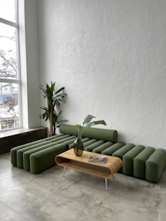 A modern living room with a green modular couch featuring cylindrical cushions and a sleek wooden coffee table with a vase containing leaves. The room has a tall potted plant in the corner and large windows allowing natural light to illuminate the textured white walls and concrete floor.