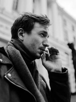 A black and white photograph of a person smoking a cigarette. They are wearing a scarf and a jacket with buttons. The background appears to be an architectural structure with tall columns, slightly out of focus.