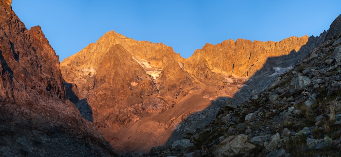 A breathtaking mountain landscape captured during sunset, where the peaks are bathed in a warm golden light. The clear blue sky contrasts with the rocky terrain, adding depth and drama to the scene.