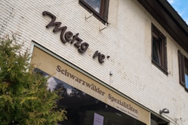 The image captures a section of a building with a light-colored brick facade. A sign with the partial name 'Metzg re' is prominently mounted on the wall in dark, cursive letters. Below this, there is a smaller brown awning with the text 'Schwarzw&auml;lder Spezialit&auml;ten aus eigener Herstellung,' which suggests a focus on specialized products. A bush with green foliage is visible in the foreground on the left side.