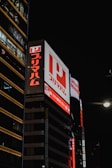 Large illuminated billboards with Japanese characters are displayed on the side of a tall building at night. Bright red and white lights are prominent with a dark sky backdrop, creating a vibrant contrast.