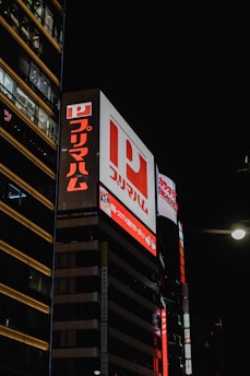 Large illuminated billboards with Japanese characters are displayed on the side of a tall building at night. Bright red and white lights are prominent with a dark sky backdrop, creating a vibrant contrast.