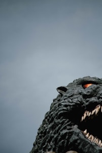 A large, detailed sculpture of a monstrous creature with rough, textured skin, sharp teeth, and an intense gaze. The creature occupies the right side, with a mostly clear sky as the backdrop.