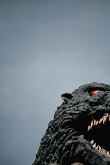 A large, detailed sculpture of a monstrous creature with rough, textured skin, sharp teeth, and an intense gaze. The creature occupies the right side, with a mostly clear sky as the backdrop.