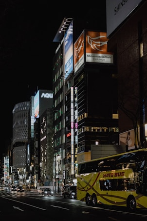 A nighttime urban street scene with brightly lit commercial buildings featuring various advertisements and signage. A yellow double-decker bus labeled 'Hato Bus' travels down the road alongside several cars. The architecture includes modern high-rise buildings illuminated by streetlights and neon signs. The atmosphere gives a bustling and lively city vibe.