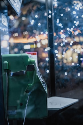 A green public phone is visible with its handset resting on the hook. The surrounding atmosphere is filled with bokeh lights of various colors, creating a dreamy and nostalgic ambiance.