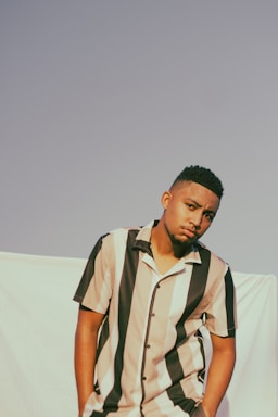 A person is leaning slightly forward against a light-colored backdrop, wearing a short-sleeved shirt with vertical black and beige stripes. The expression is neutral, and the sky is clear.