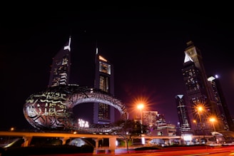 A nighttime cityscape featuring modern skyscrapers illuminated against the dark sky. A uniquely designed building with illuminated Arabic calligraphy is prominently displayed in the foreground. Streetlights cast a warm glow, while light trails hint at moving vehicles.