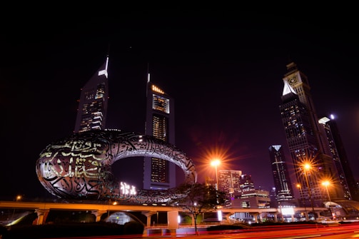 A nighttime cityscape featuring modern skyscrapers illuminated against the dark sky. A uniquely designed building with illuminated Arabic calligraphy is prominently displayed in the foreground. Streetlights cast a warm glow, while light trails hint at moving vehicles.