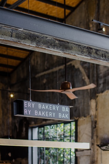 An industrial-style interior with visible steel beams and a rustic ceiling. A digital sign hanging from the ceiling displays the words 'BAKERY' repeatedly. A modern ceiling fan with wooden blades is overhead, and a large window in the background allows natural light to filter in, casting soft shadows.