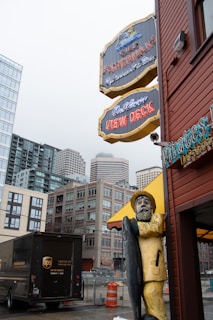A wooden statue of a fisherman holding a large fish stands near a street corner. Above, there are vintage-style signs for a restaurant and view decks. The background includes tall, modern buildings and a UPS delivery truck parked nearby. An overcast sky adds to the urban atmosphere.