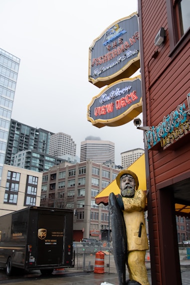 A wooden statue of a fisherman holding a large fish stands near a street corner. Above, there are vintage-style signs for a restaurant and view decks. The background includes tall, modern buildings and a UPS delivery truck parked nearby. An overcast sky adds to the urban atmosphere.