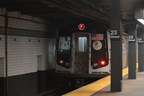 A subway train is arriving at the 23 Street station. The train is marked with an 'F' line sign and displays the American flag and MTA New York City Subway logo. The station platform has yellow safety markings and is empty. The walls are tiled with white tiles and a green stripe.