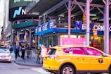 A busy urban street scene featuring the New York Police Department building with people walking on the sidewalk and a yellow taxi prominently in the foreground. Billboards and signs are visible, including a large Nasdaq advertisement.