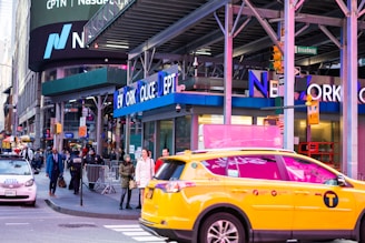A busy urban street scene featuring the New York Police Department building with people walking on the sidewalk and a yellow taxi prominently in the foreground. Billboards and signs are visible, including a large Nasdaq advertisement.
