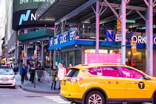 A busy urban street scene featuring the New York Police Department building with people walking on the sidewalk and a yellow taxi prominently in the foreground. Billboards and signs are visible, including a large Nasdaq advertisement.