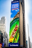 A large outdoor advertisement on a skyscraper depicts a family enjoying a water slide. The text on the advertisement reads 'Visit Orlando' and 'Anything is possible if you can imagine it.' The colors are vibrant, including a bright blue sky and a green water slide.