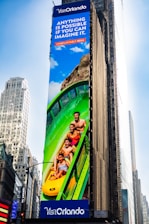 A large outdoor advertisement on a skyscraper depicts a family enjoying a water slide. The text on the advertisement reads 'Visit Orlando' and 'Anything is possible if you can imagine it.' The colors are vibrant, including a bright blue sky and a green water slide.