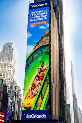 A large outdoor advertisement on a skyscraper depicts a family enjoying a water slide. The text on the advertisement reads 'Visit Orlando' and 'Anything is possible if you can imagine it.' The colors are vibrant, including a bright blue sky and a green water slide.