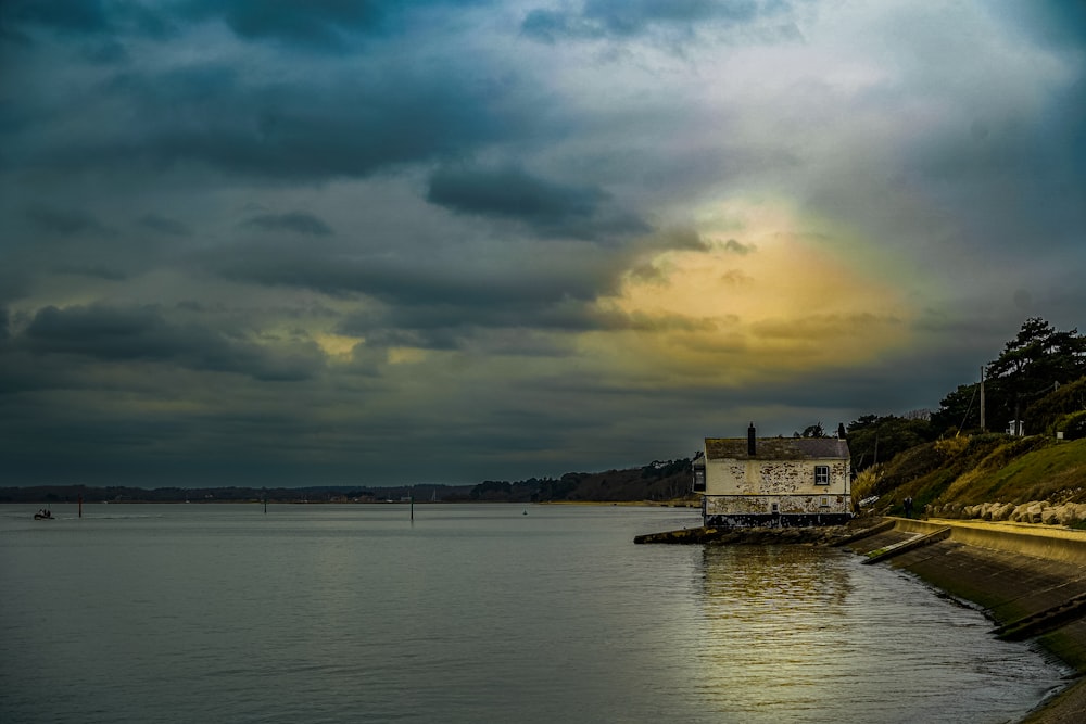a house sitting on the edge of a body of water under a cloudy sky