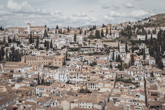 Many tightly packed, white and tan buildings with tiled roofs spread across a hilly landscape. A mix of greenery with numerous tall cypress trees is interspersed among the structures, giving a Mediterranean feel. The sky is lightly clouded, adding a serene atmosphere to the scene.