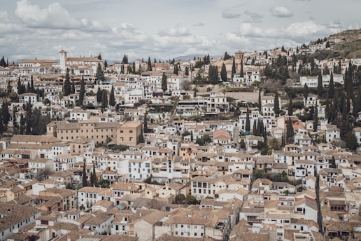Many tightly packed, white and tan buildings with tiled roofs spread across a hilly landscape. A mix of greenery with numerous tall cypress trees is interspersed among the structures, giving a Mediterranean feel. The sky is lightly clouded, adding a serene atmosphere to the scene.