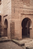 A section of an aged brick building featuring a series of geometric archways and architectural details in a historic or ancient style. The brickwork appears weathered and textured, with some areas showing signs of deterioration. The ground is paved with stone tiles, and there is a low brick wall in the foreground.