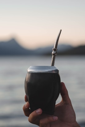 A hand holds a traditional mate cup with a metal straw against a backdrop of a serene body of water and blurred mountains in the distance. The lighting is soft, suggesting either early morning or evening.