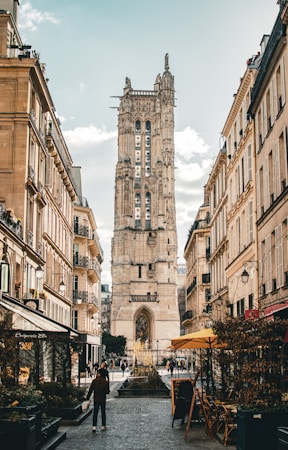 A tall, historic stone tower is situated at the center between two narrow city streets lined with classic Parisian buildings. The scene includes a few pedestrians walking, with street cafes and shops on the ground floors of the buildings. The sky is partly cloudy, contributing to a warm, inviting atmosphere.
