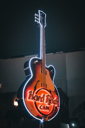A large neon sign shaped like an electric guitar is prominently displayed against a dark background. The sign features glowing red letters spelling 'Hard Rock Café,' with bright outlines surrounding the guitar shape. The lighting creates a vivid contrast with the night sky.