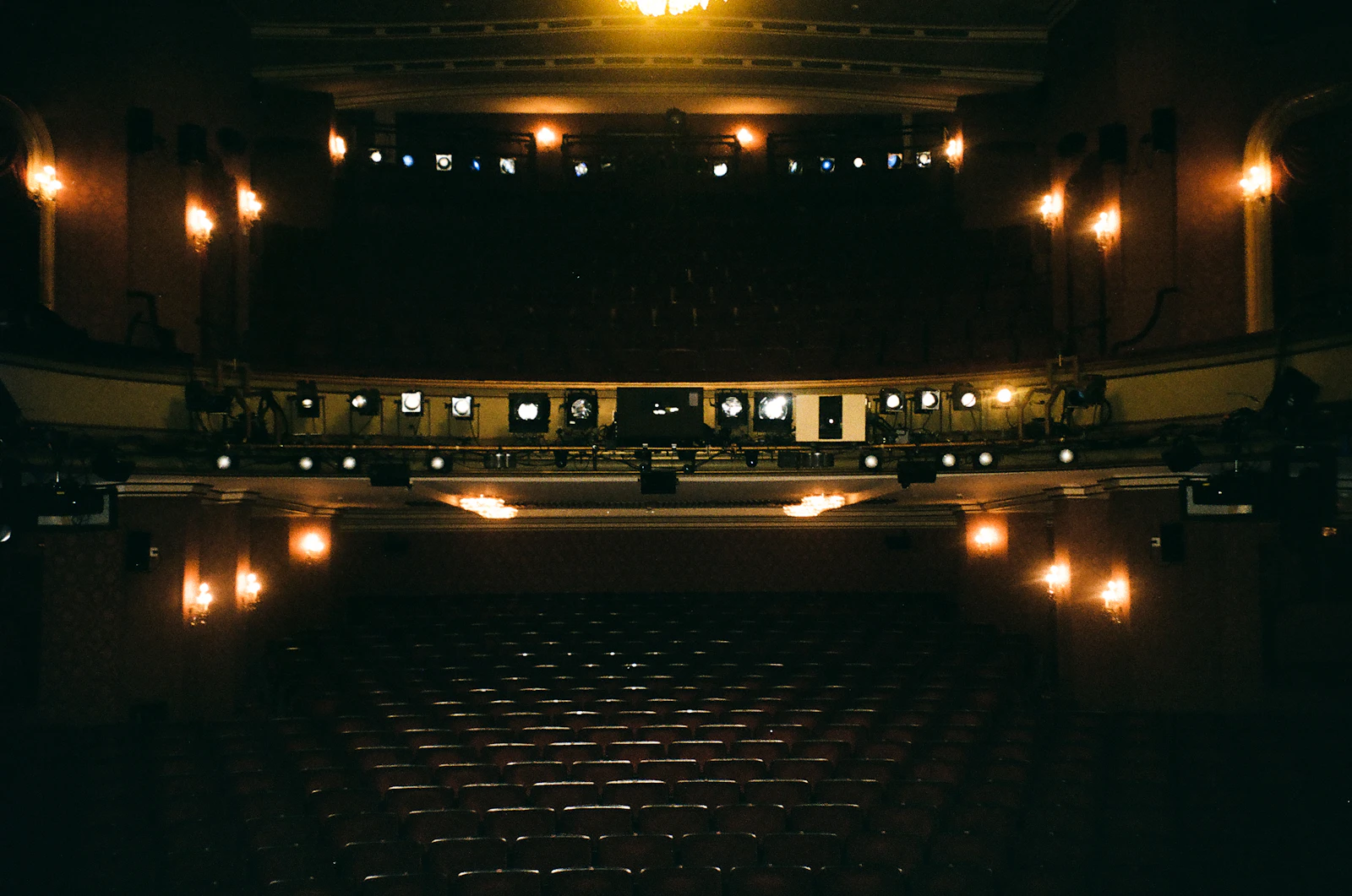 Empty theatre auditorium in near darkness, rows of seats facing a stage under a rig of warm spotlights, shot on film
