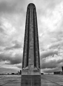 A tall, cylindrical stone monument towers against a dramatic cloud-filled sky. The surface of the monument is composed of rectangular stone blocks, with subtle textures visible. The ground is a shiny tile or stone surface that reflects the base of the monument. In the background, the silhouettes of city buildings can be seen on the horizon.