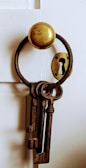 A set of old, rusty skeleton keys hangs from a round metal hook on a white wooden door. Beside them, an antique keyhole plate is visible.