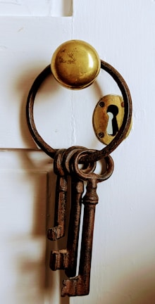 A set of old, rusty skeleton keys hangs from a round metal hook on a white wooden door. Beside them, an antique keyhole plate is visible.