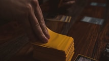 A hand is holding a stack of playing cards with yellow sleeves on a wooden table. Several cards are laid out in the background, and a dice is visible to the side.
