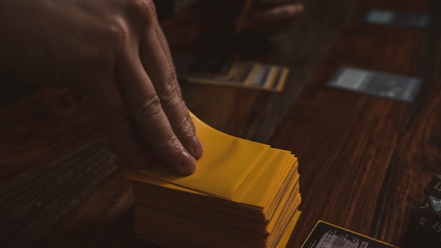 A hand is holding a stack of playing cards with yellow sleeves on a wooden table. Several cards are laid out in the background, and a dice is visible to the side.