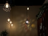 Four hanging light fixtures are displayed against a dimly lit wall. Three of these lights have a geometric metal frame with visible bulbs, emitting a warm glow. In the center, a colorful stained glass lamp casts intricate light patterns on the wall. A leafy green plant and shelves filled with bottles are visible in the background.