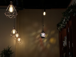 Four hanging light fixtures are displayed against a dimly lit wall. Three of these lights have a geometric metal frame with visible bulbs, emitting a warm glow. In the center, a colorful stained glass lamp casts intricate light patterns on the wall. A leafy green plant and shelves filled with bottles are visible in the background.