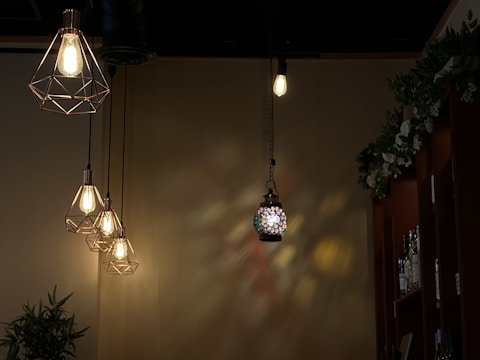Four hanging light fixtures are displayed against a dimly lit wall. Three of these lights have a geometric metal frame with visible bulbs, emitting a warm glow. In the center, a colorful stained glass lamp casts intricate light patterns on the wall. A leafy green plant and shelves filled with bottles are visible in the background.