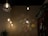 Four hanging light fixtures are displayed against a dimly lit wall. Three of these lights have a geometric metal frame with visible bulbs, emitting a warm glow. In the center, a colorful stained glass lamp casts intricate light patterns on the wall. A leafy green plant and shelves filled with bottles are visible in the background.