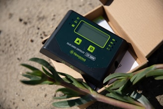 A solar charge controller is placed on a sandy surface, partially inside an open cardboard box. The device is black with green accents, featuring a small display and several buttons. A plant with green leaves is visible in the foreground, adding contrast to the industrial appearance of the controller.