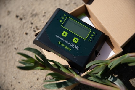 A solar charge controller is placed on a sandy surface, partially inside an open cardboard box. The device is black with green accents, featuring a small display and several buttons. A plant with green leaves is visible in the foreground, adding contrast to the industrial appearance of the controller.