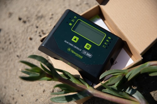 A solar charge controller is placed on a sandy surface, partially inside an open cardboard box. The device is black with green accents, featuring a small display and several buttons. A plant with green leaves is visible in the foreground, adding contrast to the industrial appearance of the controller.