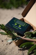 A solar charge controller device is placed inside an open cardboard box. The device has a digital screen with buttons for navigation and configuration. It is branded with 'Newpowa' and '12V/30W'. The setting appears to be outdoors, as the box is on sandy ground with green plants around it.