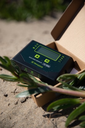 A solar charge controller device is placed inside an open cardboard box. The device has a digital screen with buttons for navigation and configuration. It is branded with 'Newpowa' and '12V/30W'. The setting appears to be outdoors, as the box is on sandy ground with green plants around it.