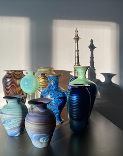 A collection of intricately designed vases and a tall candlestick are arranged on a surface. The sunlight casts strong shadows on the wall behind them, emphasizing the textures and colors of the vases. The vases have rich patterns and color variations ranging from marbled earth tones to bright blues and greens.