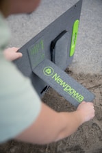 A person is setting up a solar panel with the branding 'Newpowa' visible in bright green text on a gray background. The panel is being placed on a sandy surface.